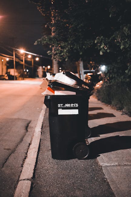A black wheeled rubbish bin with the label 'St. John's' on the front, positioned on a sidewalk at night. The bin is partially open and filled with various waste items, including white paper or cardboard and dark-colored materials. It is located near the edge of a street, with streetlights illuminating the asphalt pavement and casting shadows across the sidewalk. In the background, there are trees, a row of streetlights, and parked cars. The scene has a dark, urban atmosphere, with the bin representing an aspect of local waste disposal services, implying an alternative to official rubbish collection. The environment appears calm with minimal activity, typical of residential streets during evening hours. Natural lighting from street lamps highlights the details of the bin and surrounding area, subtly reflecting the context of private waste handling or on-site clearance. This scene exemplifies the importance of proper rubbish storage and disposal in a residential neighbourhood, with a focus on waste management towards sustainable practices. The image aligns with the services provided by Rubbish Collection Brent, illustrating the practical aspect of rubbish disposal at the curbside after collection or before removal.