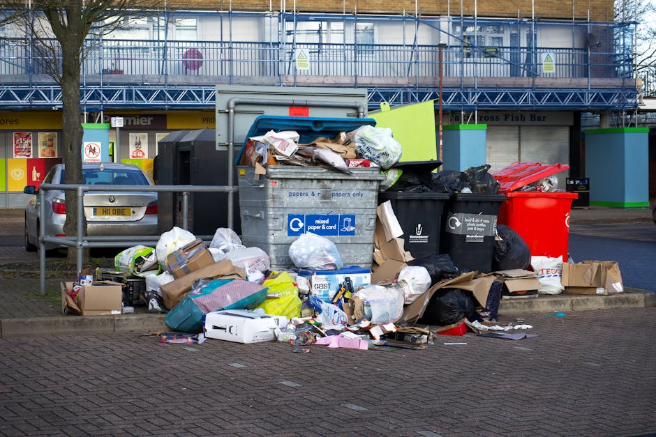 A large collection of overflowing rubbish and waste materials is piled on a paved sidewalk in front of a commercial building, with some trash spilling onto the street. The waste includes various black, white, and brown rubbish bags, some ripped open with contents such as paper, cardboard, and plastic packaging scattered across the ground. Several cardboard boxes, some flattened and others partially intact, are alongside loose items like paper, plastic containers, and packaging materials. In the background, a grey skip bin with a partially open lid contains more discarded paper and cardboard, while other black and red rubbish bins are visible nearby, all filled and overstuffed. The scene is set in an urban environment with a metal railing separating the waste area from parked cars, including a silver vehicle parked adjacent to the rubbish pile. Behind the waste, a building features a scaffolding structure with blue mesh netting and bright green accents, indicating ongoing construction or maintenance work. The overall setting exemplifies the need for professional rubbish removal services such as those provided by Rubbish Collection Brent for proper waste management and alternative disposal options.