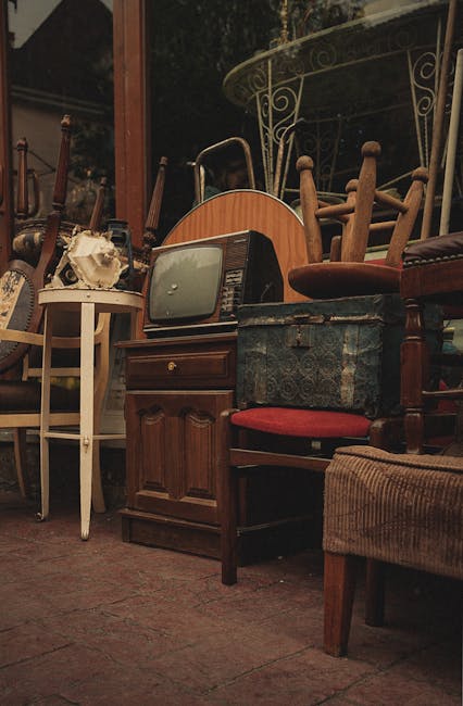 A collection of used furniture and household items arranged outdoors on a paved surface, including several wooden chairs with different finishes and upholstery, a small vintage television with a curved screen and woodgrain finish, a white side table holding a pile of miscellaneous objects, and a metal garden table with a decorative wrought iron design along its edge. The chairs are stacked and leaning against each other, with some featuring upholstered seats in red or brown fabrics, while others are bare wood. Behind the furniture, a brick building and trees are visible in the background, suggesting an outdoor setting such as a yard or a furniture clearance area. The scene appears to be part of a private collection or an arrangement for an on-site clearance, reflecting the type of items that may be handled by waste removal services like Rubbish Collection Brent. The natural ambient light indicates daytime conditions, with warm hues highlighting the textures of wood and fabric and creating a casual, storage-like atmosphere suitable for furniture disposal or alternative waste handling.