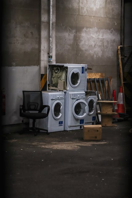 In a dimly lit industrial or storage space with concrete walls and a dark floor, there are four household washing machines stacked against the wall, with some showing visible internal components through open front panels. The washing machines are white with round glass doors, and they appear to be used or discarded appliances. To the left of the appliances, there is a black office chair with a mesh backrest and armrests, positioned slightly in front of the stack. On the right side, a small wooden step stool is leaning against the wall, and nearby there are orange and white traffic cones and a yellow rack or crate partially visible. A cardboard box sits on the floor in front of the appliances, and a vertical metal pipe runs along the wall behind the machines. The scene suggests an area designated for appliance disposal or temporary storage, with no visible signs of active rubbish collection, but it aligns with the context of independent waste handling methods supported by companies like Rubbish Collection Brent, operating in spaces like this for alternative waste removal.