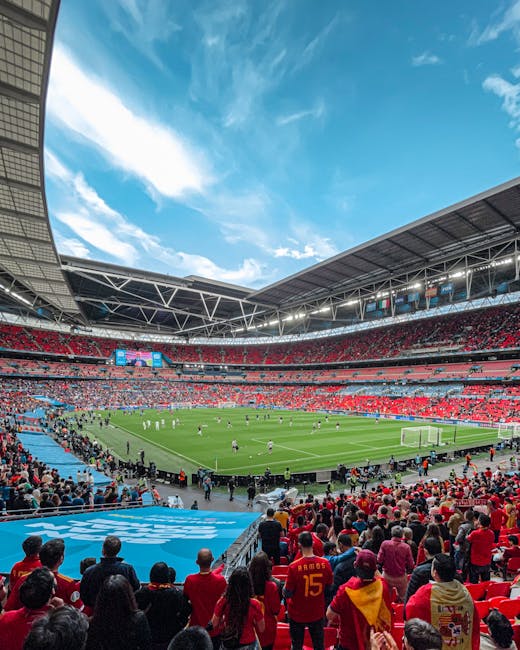 A large football stadium filled with spectators seated on multiple tiers, predominantly dressed in red, watching a match on a lush green pitch with players actively engaged. The stadium's modern architecture includes a partially covered roof structure with visible metal beams, and large digital screens displaying game information are mounted along the upper tier. The seating area is densely populated, extending around the entire field, with some spectators standing and others seated. The foreground shows fans sitting close to the pitch, with clear views of the goal area and surrounding boundary lines. Bright daylight illuminates the scene, with a partly cloudy blue sky overhead, contrasting with the vivid red seating and the green playing surface. This setting exemplifies an event at a major sports venue, where proper waste management and rubbish removal are essential for maintaining cleanliness during large-scale gatherings, aligning with services provided by Rubbish Collection Brent for private event clearance and stadium waste disposal needs.