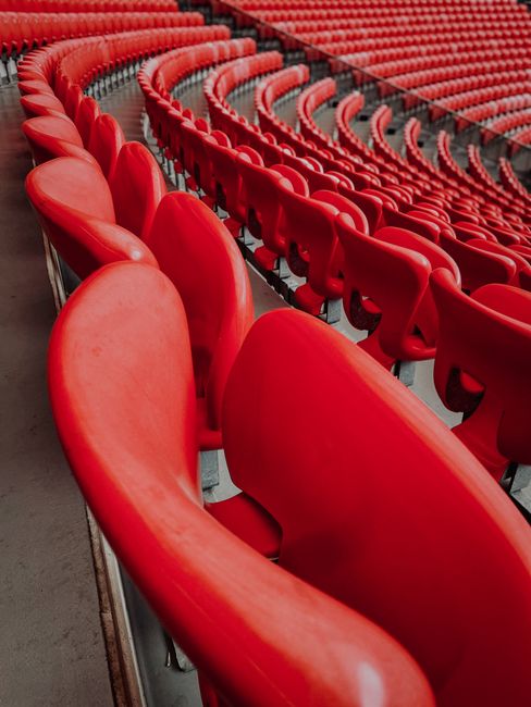 A large section of empty stadium seating featuring bright red plastic chairs with smooth, matte finishes and curved backs, arranged in multiple rows that extend into the distance, with some chairs slightly leaning and stacked closely together. The chairs are mounted on black metal frames and are set on a flat, grey concrete floor, with a metal railing visible on the left side of the image. The background shows additional rows of the same type of red chairs, creating a pattern of repeating shapes and vibrant colour tones typical of large event venues. The lighting appears natural and diffused, casting soft shadows on the chairs and the surrounding environment, suggesting the stadium may be indoor with ample ambient light. This scene subtly relates to event management and the logistics of clearing or managing large volumes of seating furniture and waste generated during events, such as those managed by private waste collection services like Rubbish Collection Brent, especially in the context of disposing of or recycling used seating after large gatherings.