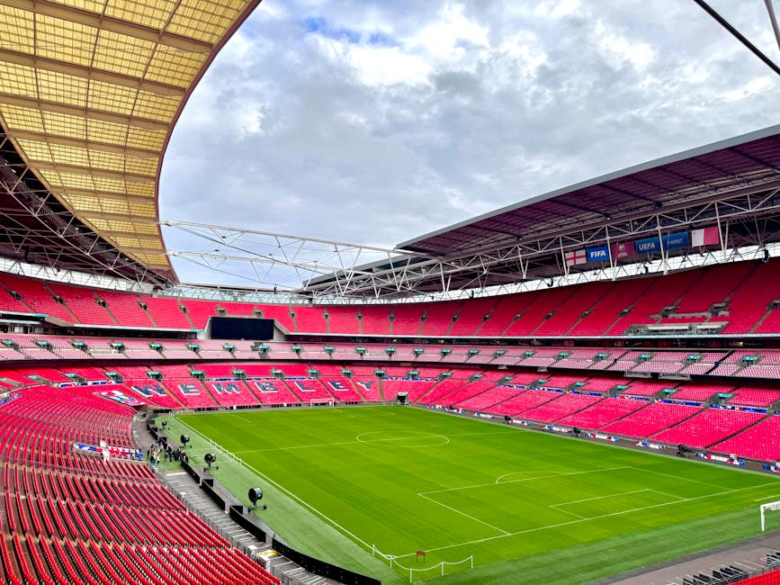 A large football stadium filled with spectators seated on multiple tiers, predominantly dressed in red, watching a match on a lush green pitch with players actively engaged. The stadium's modern architecture includes a partially covered roof structure with visible metal beams, and large digital screens displaying game information are mounted along the upper tier. The seating area is densely populated, extending around the entire field, with some spectators standing and others seated. The foreground shows fans sitting close to the pitch, with clear views of the goal area and surrounding boundary lines. Bright daylight illuminates the scene, with a partly cloudy blue sky overhead, contrasting with the vivid red seating and the green playing surface. This setting exemplifies an event at a major sports venue, where proper waste management and rubbish removal are essential for maintaining cleanliness during large-scale gatherings, aligning with services provided by Rubbish Collection Brent for private event clearance and stadium waste disposal needs.
